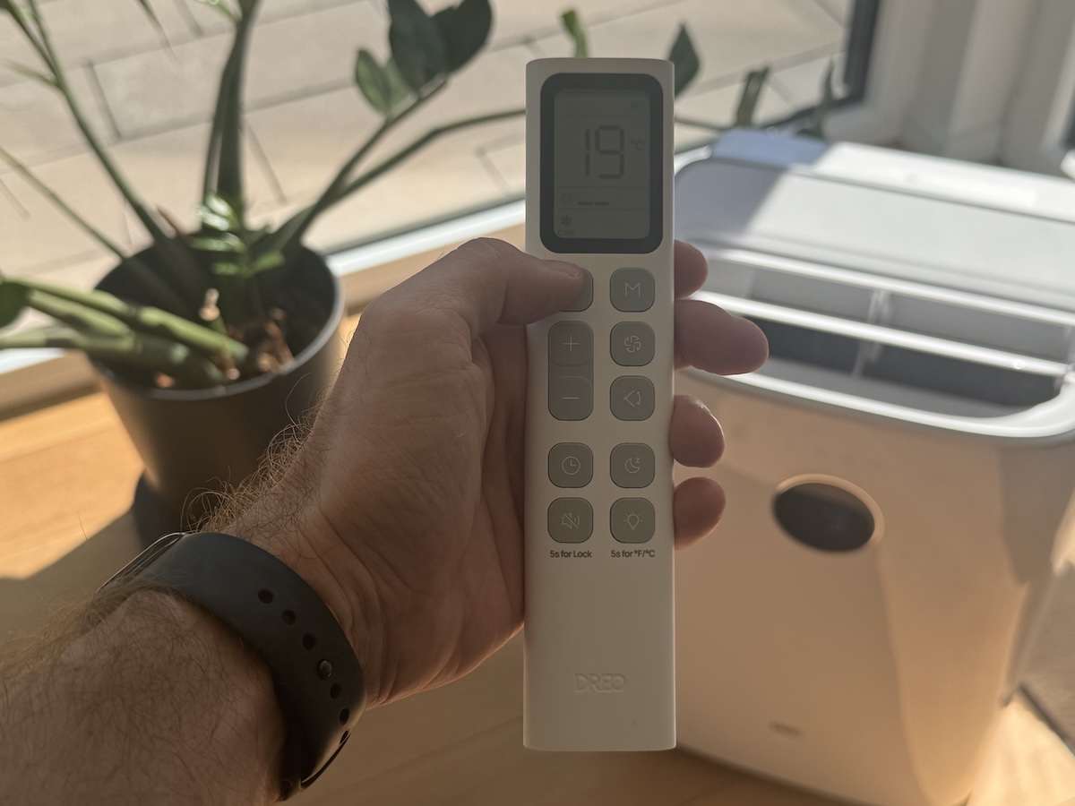 Fernbedienung der Dreo Mobile Klimaanlage 318S mit großem Display und großen Tasten in der Hand, im Hintergrund das Gerät am Fenster