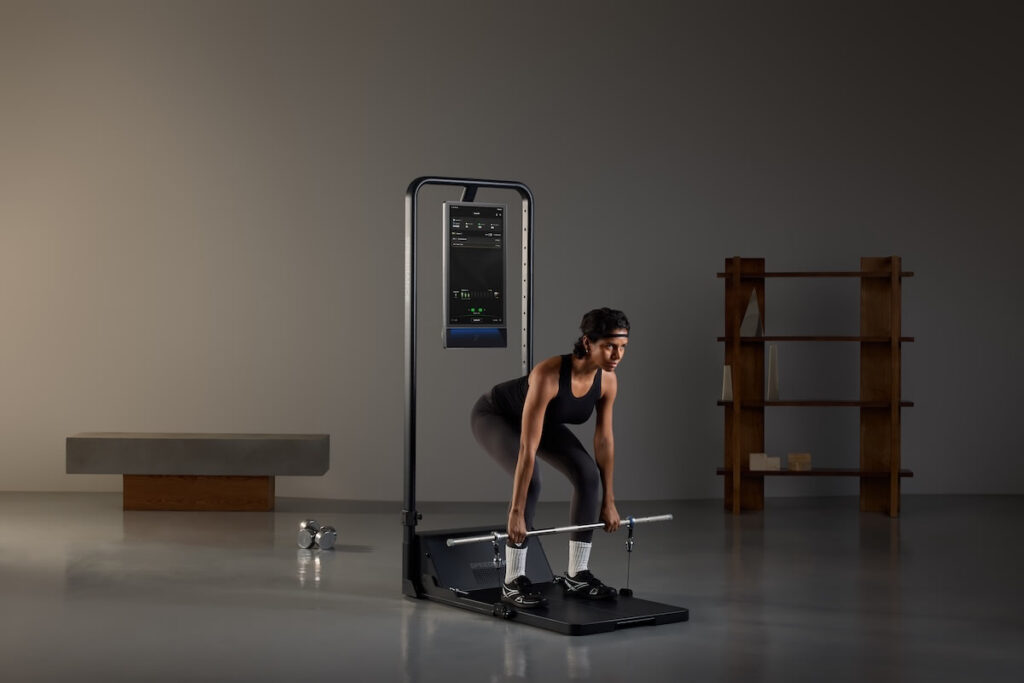 Eine Frau trainiert Kreuzheben mit dem Speediance Gym Monster 3, einem kompakten Smart-Fitnessgerät mit digitalem Display und Langhantel-Aufsatz, in einem minimalistisch eingerichteten Raum.