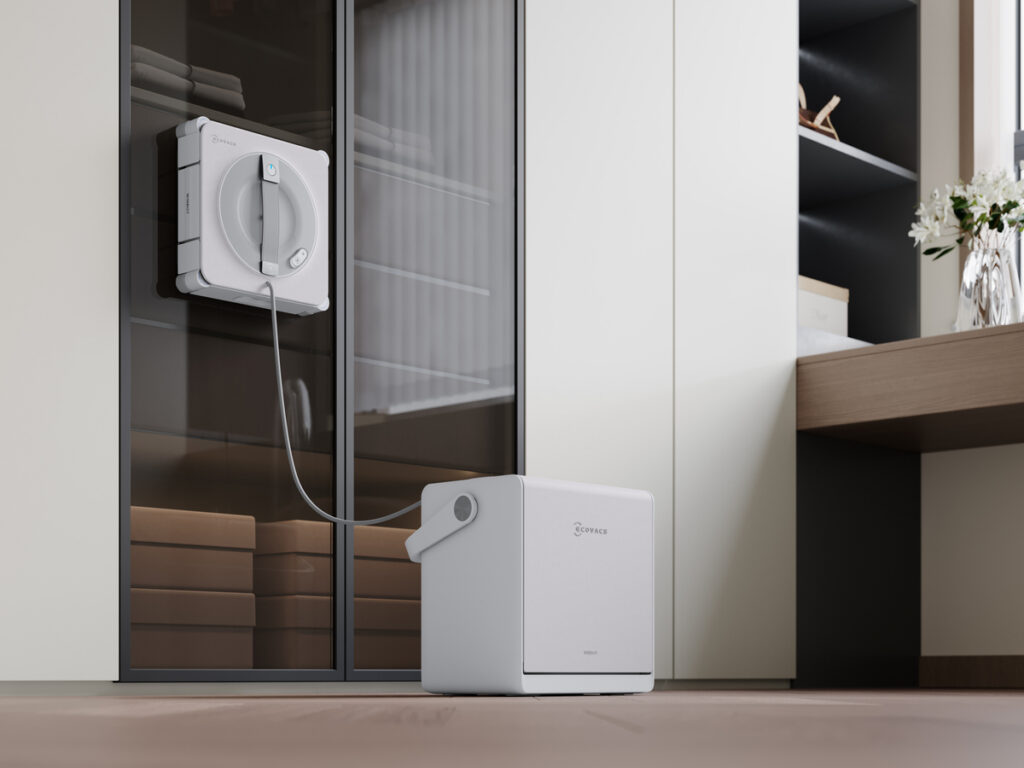 Weißer Ecovacs Winbot W2S Pro Omni Fensterputzroboter an einer Glastür befestigt, verbunden mit einer würfelförmigen Station am Boden.