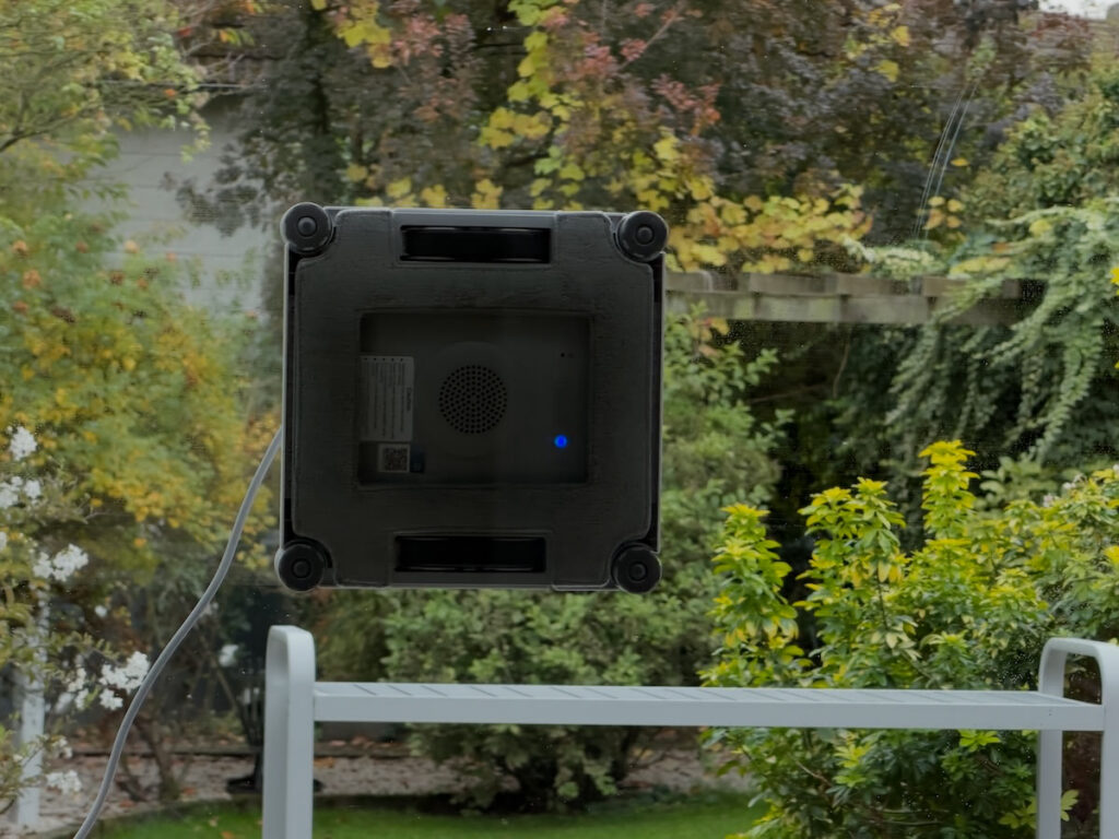 Ecovacs Winbot W2S Omni im Einsatz: Der Fensterputzroboter haftet an einer großen Glasscheibe, dahinter Garten und Geländer