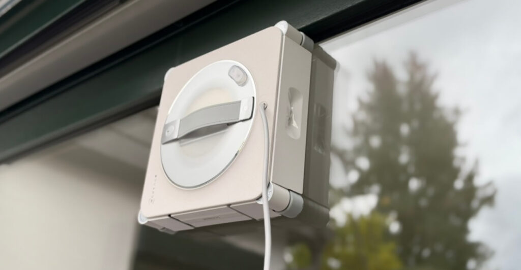 Ecovacs Winbot W2S Omni im Test: Der Fensterputzroboter haftet an einer Fensterscheibe, dahinter Bäume im Garten